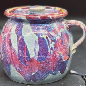 May include: A handmade ceramic pot with a lid, featuring a vibrant blend of colors. The pot showcases a marbled effect with shades of red, purple, blue, and cream. It has a handle and a matching lid, perfect for kitchen or home decor.