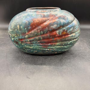 May include: A ceramic vase with a round shape and a wide opening. The vase features a textured surface with a swirl design. The colors include shades of teal, red, and yellow.