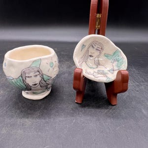 May include: Two handmade ceramic bowls with a white glaze and hand-painted designs. The larger bowl features a woman with long hair and a mountain scene. The smaller bowl features a woman with long hair and a mermaid tail.