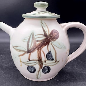 May include: A ceramic teapot with a lid, spout, and handle. The teapot is light pink with a painted design of brown branches, green leaves, and dark blue fruit. The lid and handle are light green.