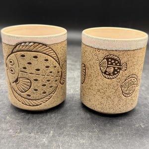 May include: Two cylindrical ceramic cups with a speckled tan finish and a light pink rim. The left cup features a large, detailed flatfish illustration, while the right cup displays two smaller fish with bubbles, all rendered in dark brown etched lines.
