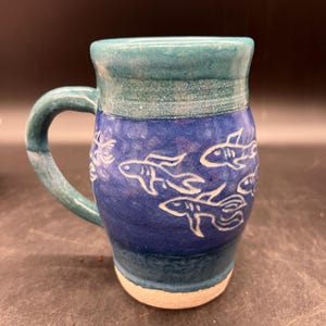 May include: A blue and green ceramic mug with a white design of fish swimming. The mug has a handle and is glazed.