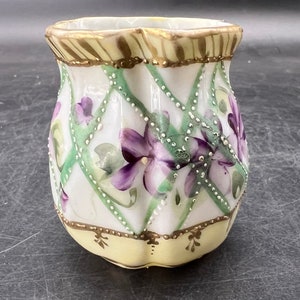 May include: A small, white ceramic pitcher with a yellow base and a floral design. The pitcher is decorated with purple and green flowers and gold accents.
