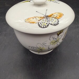 May include: A white ceramic bowl with a lid, adorned with a butterfly and floral design. The butterfly features orange, yellow, and black details. The bowl has a rounded shape and is decorated with delicate white and yellow flowers.