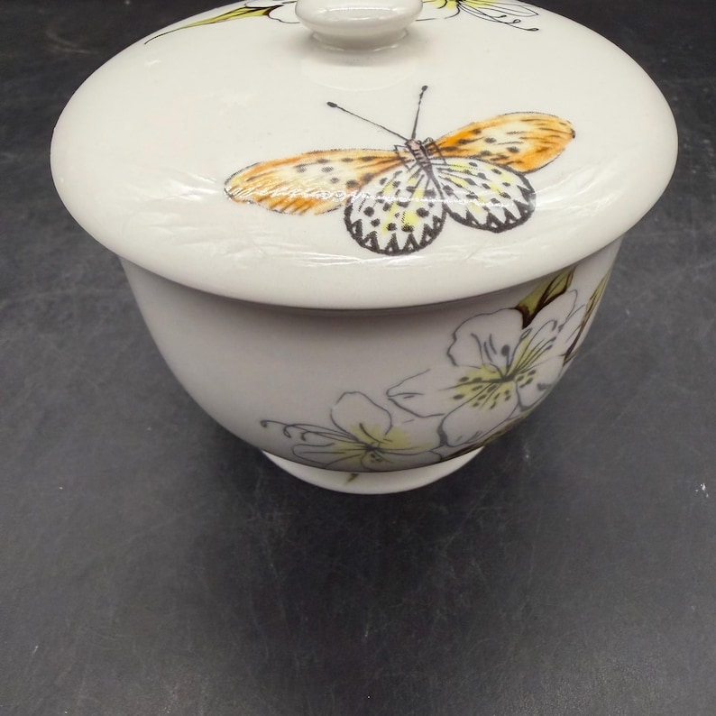 May include: A white ceramic bowl with a lid, adorned with a butterfly and floral design. The butterfly features orange, yellow, and black details. The bowl has a rounded shape and is decorated with delicate white and yellow flowers.
