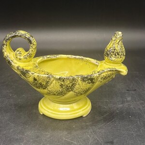 May include: A vintage ceramic oil lamp in a vibrant yellow hue with speckled black accents. The lamp features a circular handle, a spout, and a decorative finial. It is mounted on a round pedestal base, showcasing a retro design.