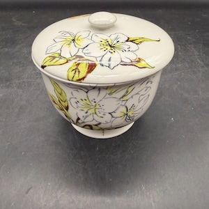 May include: A white ceramic bowl with a lid, adorned with a floral design. The flowers are white with yellow and brown accents, and the bowl has a rounded shape with a small base. The lid has a small knob.