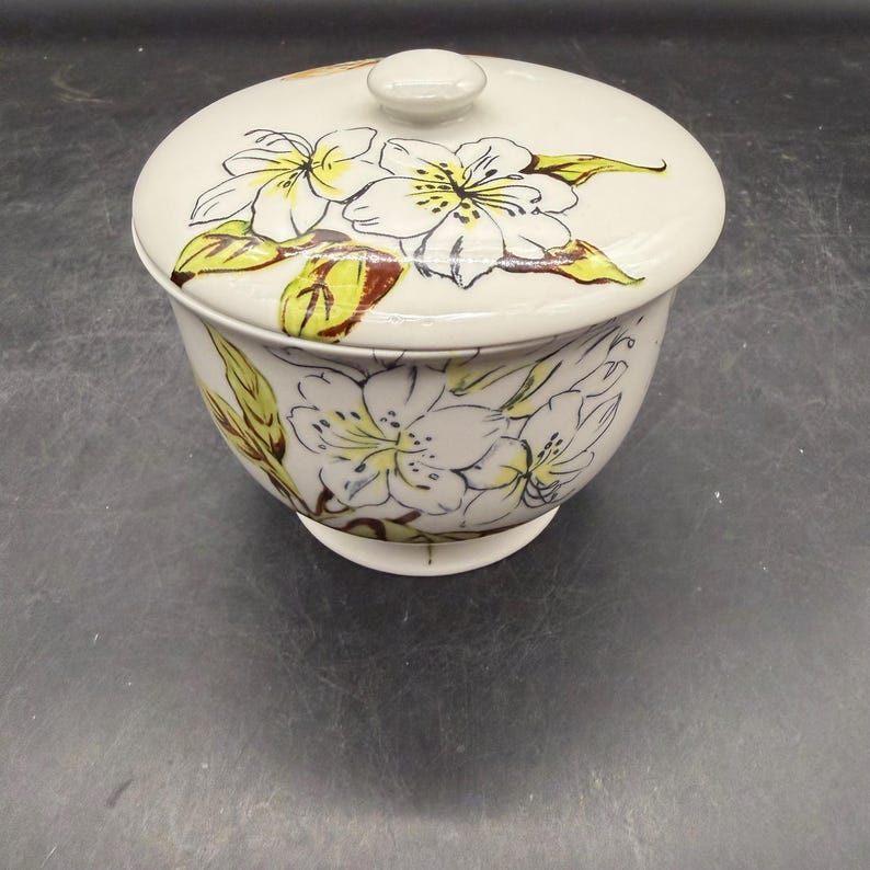 May include: A white ceramic bowl with a lid, adorned with a floral design. The flowers are white with yellow and brown accents, and the bowl has a rounded shape with a small base. The lid has a small knob.