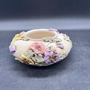 May include: A white ceramic planter with a raised design of purple, pink, and yellow pansies. The planter is shaped like a bowl and has a wide opening.