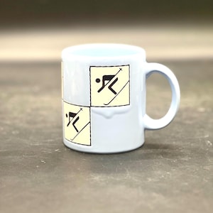 May include: A light blue ceramic mug with a white and black skier graphic. The graphic is repeated on the mug.