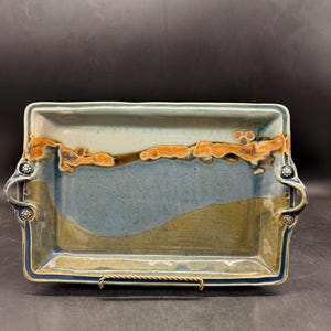 May include: Rectangular ceramic serving tray with a blue, brown, and tan color scheme. The tray has handles on each side and decorative floral accents. The rim is a darker blue, and the tray is displayed on a metal stand.
