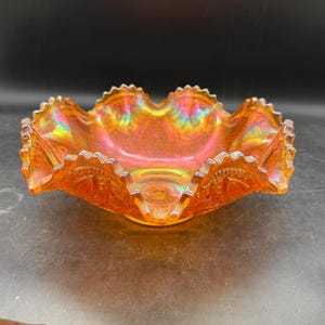May include: An iridescent orange glass bowl with a ruffled edge. The bowl has a detailed pattern around the rim and a textured interior. The colors shift from orange to yellow and green.