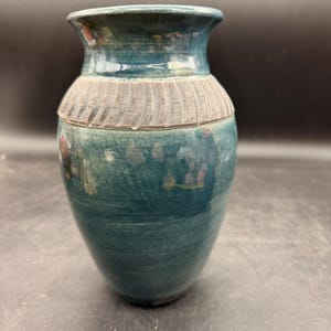 May include: A teal ceramic vase with a textured, ridged band around the middle. The vase has a flared top and a rounded body. The surface has a mottled appearance with hints of other colors.