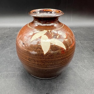 May include: A brown ceramic vase with a round body and a narrow neck. The vase features a cream-colored leaf design and a dark brown rim. The vase is a decorative item.