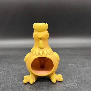 May include: A yellow ceramic chicken-shaped decorative item. The chicken has a detailed comb, wattle, and feet. It features a circular opening in the center, and is likely a decorative planter or holder. The item is set against a dark background.