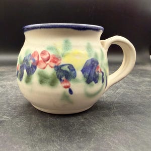 May include: Cream-colored ceramic mug with a rounded body and a handle. The mug is decorated with hand-painted flowers in red, blue, and yellow, with green leaves. The rim is trimmed with a dark blue band. A charming piece for enjoying beverages.