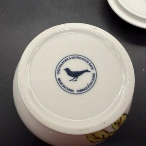 May include: White ceramic mug with a floral design and a blue bird logo on the bottom. The text "Dishwasher & Microwave Safe", "Made in China", and "Fabriqu&eacute; en Chine" are printed around the bird logo.