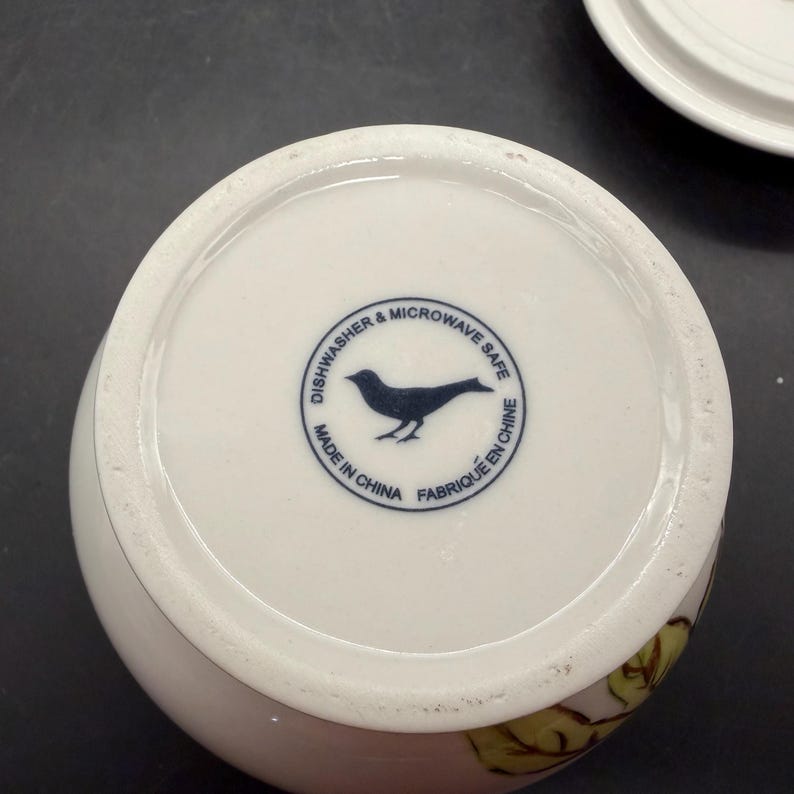 May include: White ceramic mug with a floral design and a blue bird logo on the bottom. The text "Dishwasher & Microwave Safe", "Made in China", and "Fabriqu&eacute; en Chine" are printed around the bird logo.