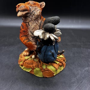 May include: A ceramic figurine of a tiger wearing a blue golf bag with a silver daisy and golf clubs. The base of the figurine is decorated with brown and green leaves. The figurine is signed "Tiger" on the base.