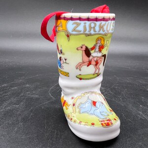 May include: A white ceramic boot ornament with a circus theme. The boot is decorated with a colourful scene of a girl riding a horse, a man playing a trumpet, and an elephant. The boot has a red ribbon attached to the top and the word "Zirkus" is written on the side.