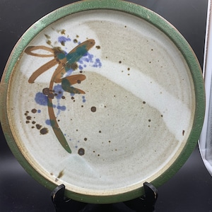 May include: A white ceramic plate with a green rim and abstract brown, blue, and green painted design. The plate has a speckled glaze.