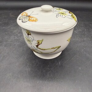 May include: A white ceramic bowl with a lid. The bowl and lid are decorated with a butterfly and floral design in yellow, orange, and green. The bowl has a pedestal base.