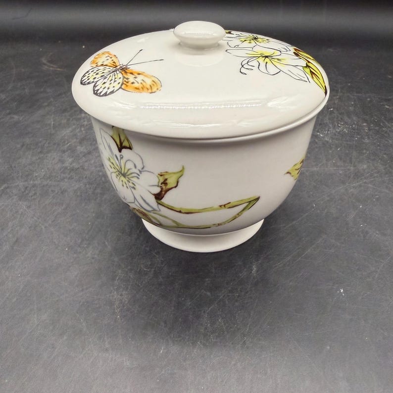 May include: A white ceramic bowl with a lid. The bowl and lid are decorated with a butterfly and floral design in yellow, orange, and green. The bowl has a pedestal base.