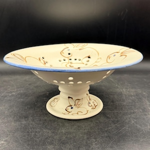 May include: A cream-colored ceramic pedestal bowl with a blue rim and brown bunny illustrations. The bowl has a perforated design around the stem and bunny illustrations on the base. The bowl is approximately 8 inches in diameter.