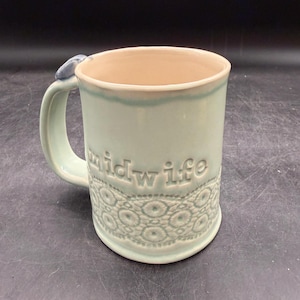 May include: A light mint green ceramic mug with a glossy finish, featuring the embossed text "midwife" on the front. The lower portion of the mug displays a raised, intricate lace-like pattern, and a small blue accent is visible on the handle.