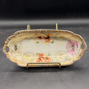 May include: Oval porcelain serving dish with a floral design. The dish features a cream-colored base with hand-painted flowers in shades of pink, orange, and white. The rim is trimmed with gold detailing and has small handles on each end.