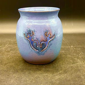 May include: A handcrafted ceramic vase with a rounded body and a flared rim. The vase is glazed in shades of blue and purple, with a deer-like design in blue and copper tones. A decorative piece for home decor.