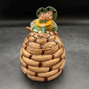 May include: A ceramic cookie jar shaped like a woven beehive with a textured brown finish. Atop the lid sit two anthropomorphic bee figures with green hats, yellow and black striped bodies, and small wings, embracing each other in a seated position.