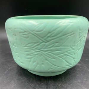 May include: A light green ceramic planter with a raised leaf design. The planter is round and has a wide opening.