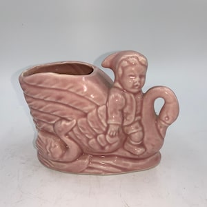 May include: A pink ceramic planter shaped like a swan with a small boy riding on its back. The planter has a small opening on the top for planting.
