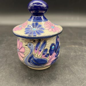 May include: A small ceramic jar with a lid, decorated with a blue and pink floral pattern and a bird design. The jar has a blue and white knob on the lid.