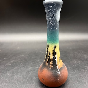 May include: A small, hand-painted ceramic vase with a night sky scene. The vase is brown with a yellow, green, and blue gradient. The scene depicts a forest with trees and a moon in the sky.