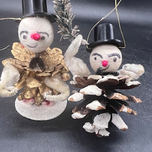 May include: Two vintage snowman ornaments made from pine cones and painted white. The snowmen are wearing black top hats and have painted faces with red noses.