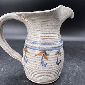 May include: A white ceramic pitcher with a brown, blue, and orange floral design. The pitcher has a wide mouth and a handle.