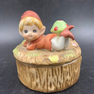 May include: A ceramic trinket box featuring a small elf figurine with a red hat and a mushroom. The elf is lying on its side with its legs outstretched. The box is decorated with a brown, tree-like design.