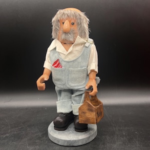 May include: A handcrafted figurine of an elderly man, wearing a white shirt, denim overalls, and black shoes. The figure has a gray beard and hair, and is holding a wooden toolbox. A red and white handkerchief is in the pocket.