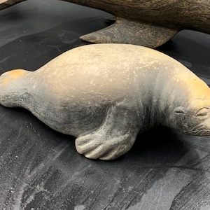 May include: A detailed sculpture of a seal, primarily gray with tan and yellow accents. The seal is lying down, with its flippers and head clearly visible. The sculpture has a textured surface, giving it a realistic appearance.