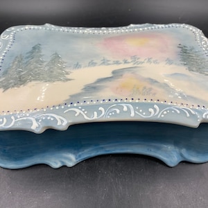 May include: A blue and white porcelain trinket box with a painted winter scene on the lid. The scene features a snowy landscape with trees and a sunset.