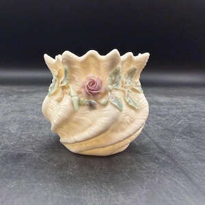 May include: A cream-colored ceramic vessel shaped like a fluted seashell with a scalloped rim. The exterior features a sculpted pink rose blossom surrounded by delicate green leaves and small blue accents, all finished with a subtle iridescent sheen.