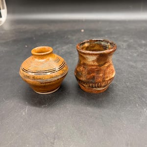 May include: Two small, handcrafted ceramic vases. One is round with horizontal black stripes and a brown glaze. The other is a small, cylindrical vase with a textured, brown and tan glaze. Both are on a dark surface.