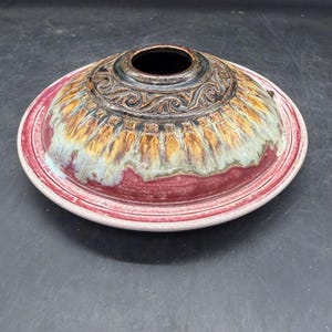 May include: A circular ceramic bowl with a wide, flat rim and a domed center featuring a central opening. The piece displays a multi-colored glaze with a deep red base, transitioning into shades of yellow, brown, and green, with carved scrollwork.