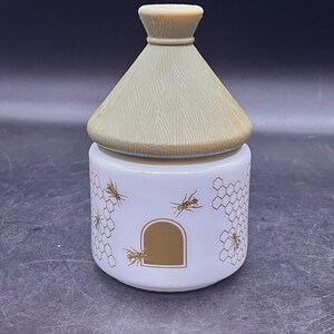 May include: A white ceramic jar with a beehive design. The jar features a brown arched doorway, honeycombs, and bee illustrations. The lid is tan and shaped like a thatched roof. A decorative kitchen item.