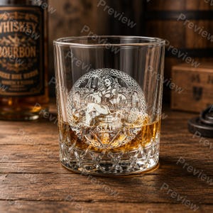 Puede incluir: Un vaso de whisky transparente con un dise&ntilde;o circular grabado de una figura sentada que sostiene una jarra, rodeada de intrincados patrones de nudos y la palabra SKAL. El vaso tiene una base pesada y facetada y contiene l&iacute;quido &aacute;mbar.
