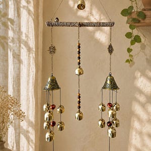 Può includere: Un carillon a vento sospeso con un ramo orizzontale testurizzato appeso a corde con una campana centrale. Due grandi campane in ottone decorate pendono dai lati, arricchite da campanellini e perline rosse, che terminano con monete metalliche circolari.