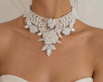 May include: A white choker necklace featuring intricate lace embroidery, floral motifs, and small white beads. The design includes a central flower element with a dangling leaf-shaped pendant, all crafted with detailed stitching and beadwork.