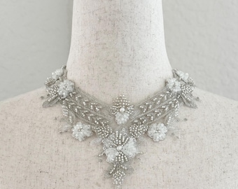 May include: A decorative collar piece featuring intricate silver metallic thread embroidery, clear glass beads, and white floral-shaped bead clusters arranged in a symmetrical pattern. The design includes leaf-like motifs and a central pointed drop.